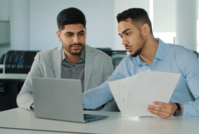 Diverse male employees Arab young colleagues discussing working together, checking emails. Two serious focused East Indian businessmen sitting in modern office using laptop looking at computer screen. High quality photo